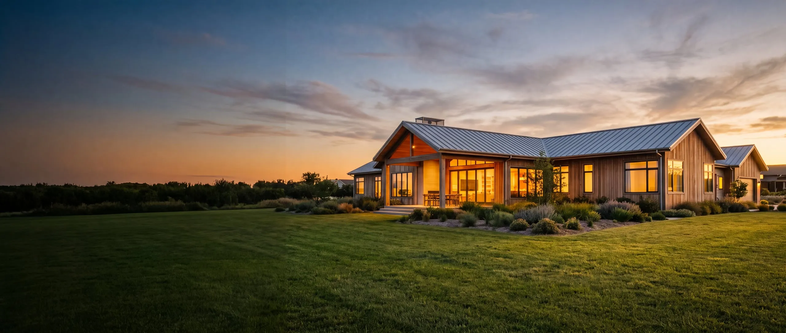 A Texas ranch home with a standing seam metal roof, warmly lit at dusk under a sunset sky
