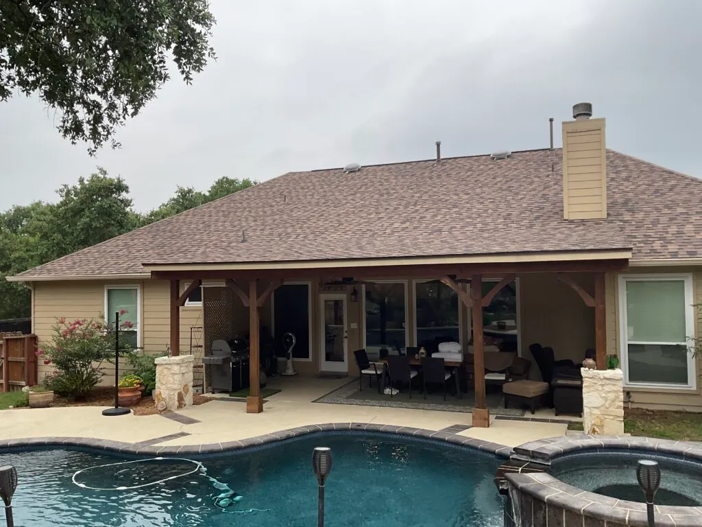 The back of a Texas home showing its shingle roof, fascia, soffit, painted siding, chimney cladding and a timber-framed covered patio